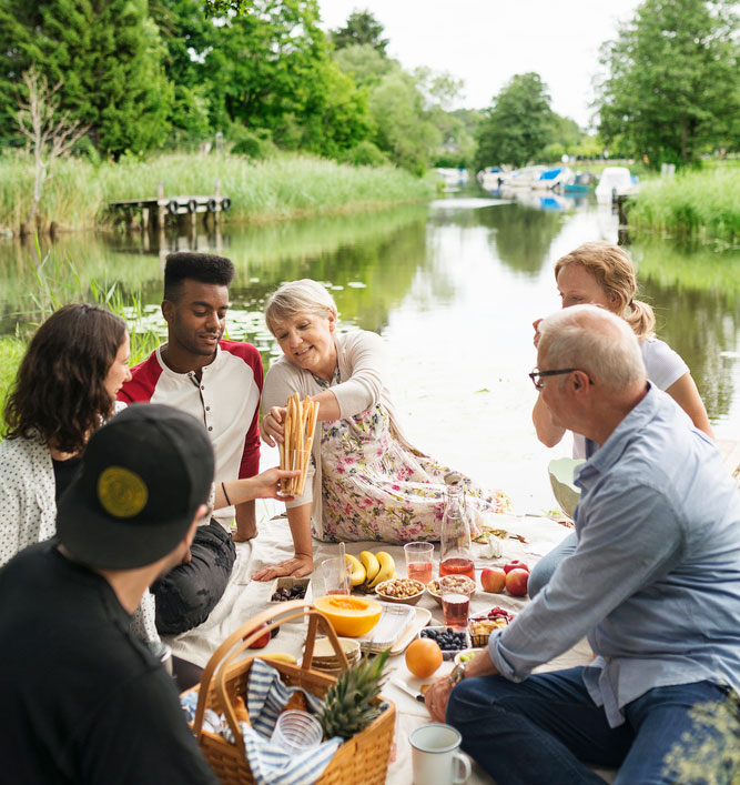 Ett sällskap med gamla och unga personer har picknick vid en å. Foto.
