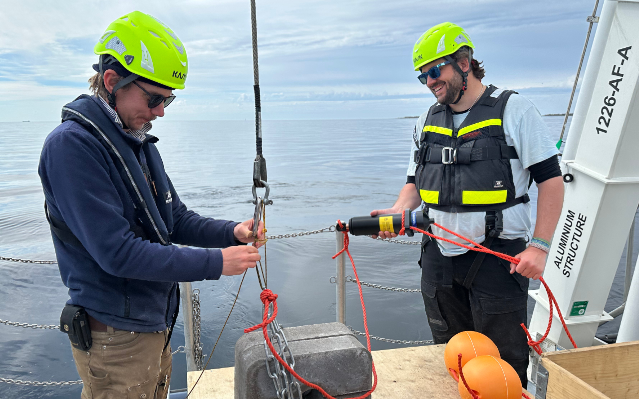 Two people in safety gear work with ropes and equipment on a boat deck over calm water, preparing scientific instruments for deployment.