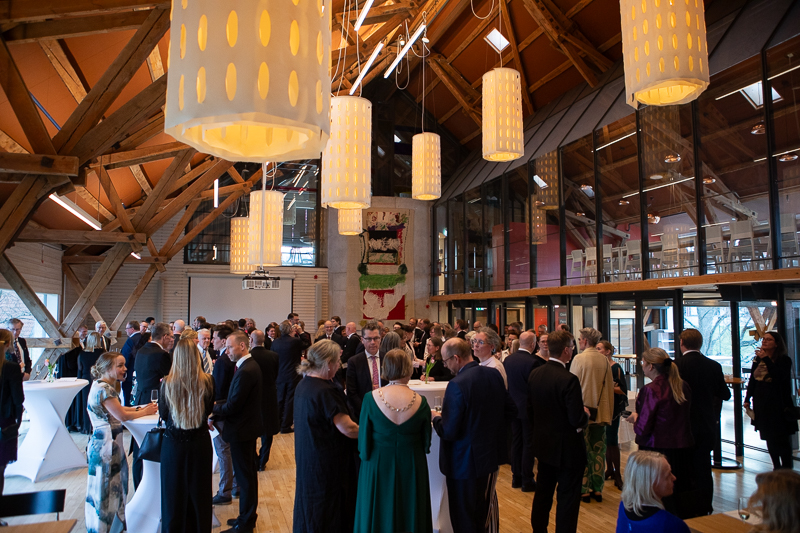 In a big hall with high ceilings, a large crowd is gathered mingling. Big white lamps hang from the ceiling.