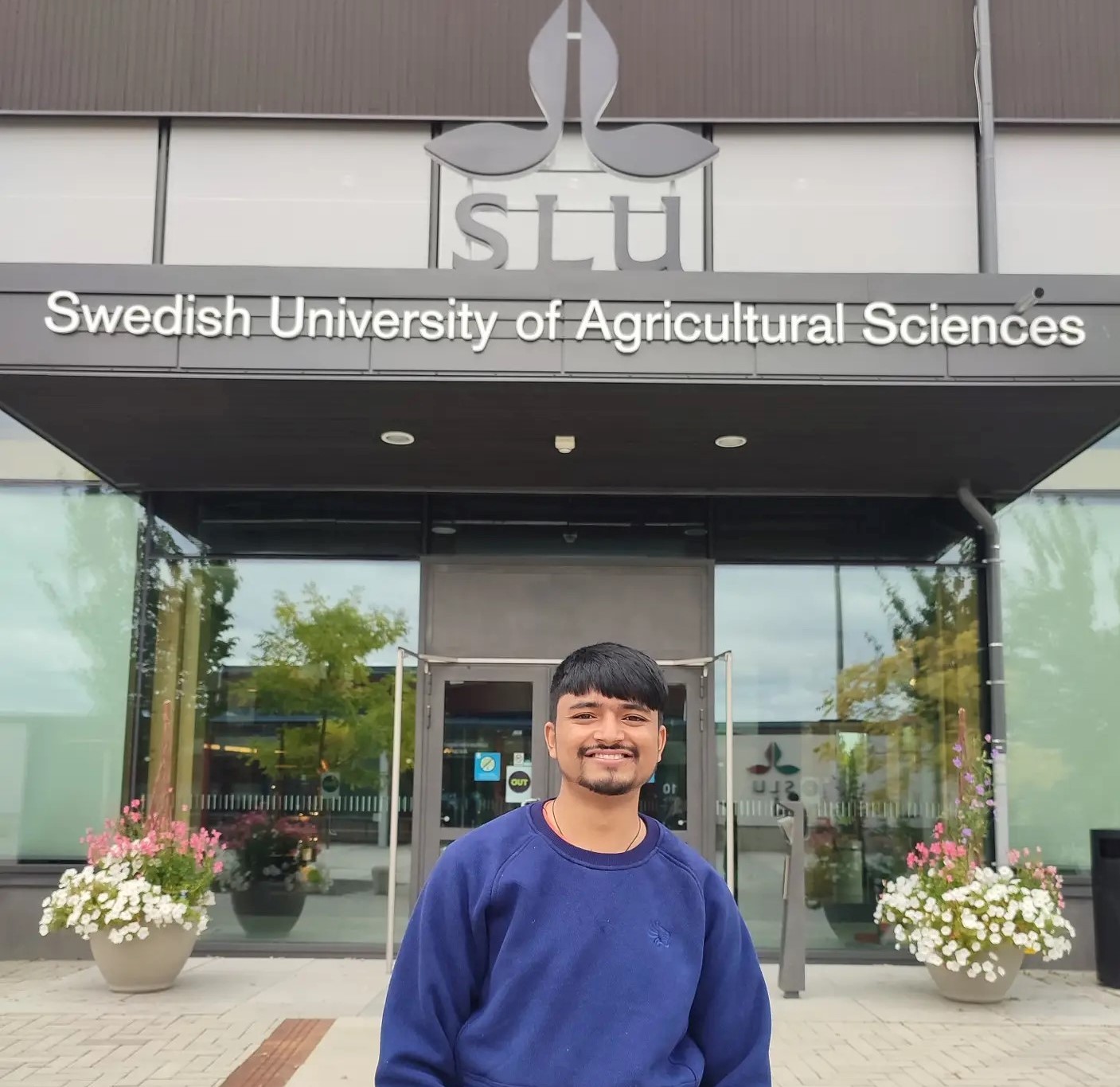 An image of a man standing in front of a house. The house has a sign that says "SLU Swedish University of Agricultural Sciences