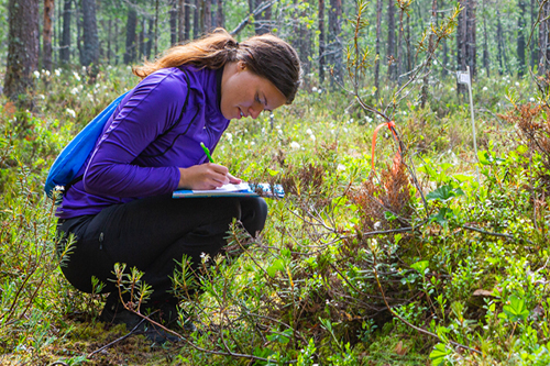 Bilden visar en student som sitter på huk i skogen och skriver på ett papper. 