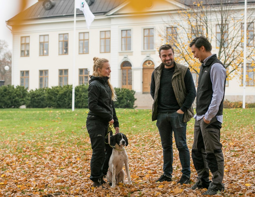 Tre studenter med en hund utanför en byggnad i Skinnskatteberg, foto.
