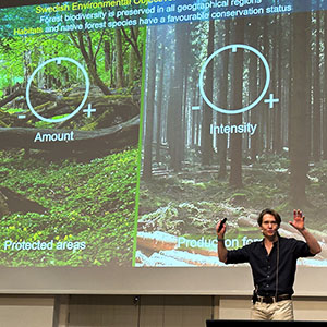 Adam Felton gestikulerar engagerat framför en bild på skog där det står Intensty