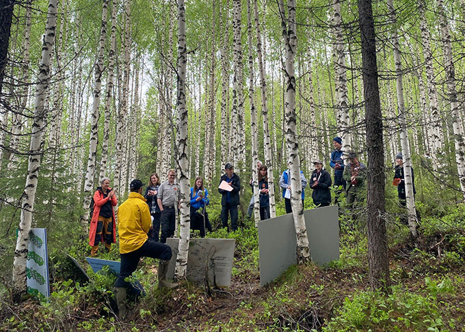 En grupp människor lyssnar till en man med postrar i en björkskog. Foto.