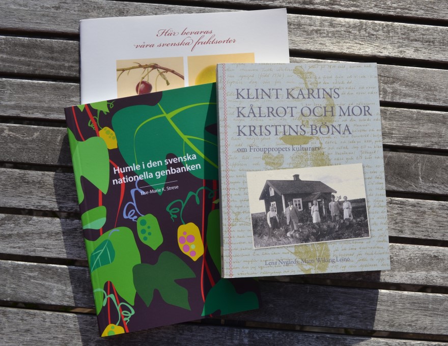 Färgfoto föreställande tre böcker som ligger på ett träbord. Böckerna är "Klint Karins kålrot och mor Kristins böna", "Humle i den svenska nationella genbanken" och "Här bevaras våra svenska fruktsorter".