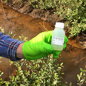 En hand med handske som håller i en provflaska ovanför ett vattendrag. Foto.