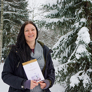 Porträttfoto av Bodil Häggström som står framför snötäckta träd, håller sin doktorsavhandling i handen och ler in i kameran. Hon har mörkt långt hår, en svart jacka och en grå tröja.