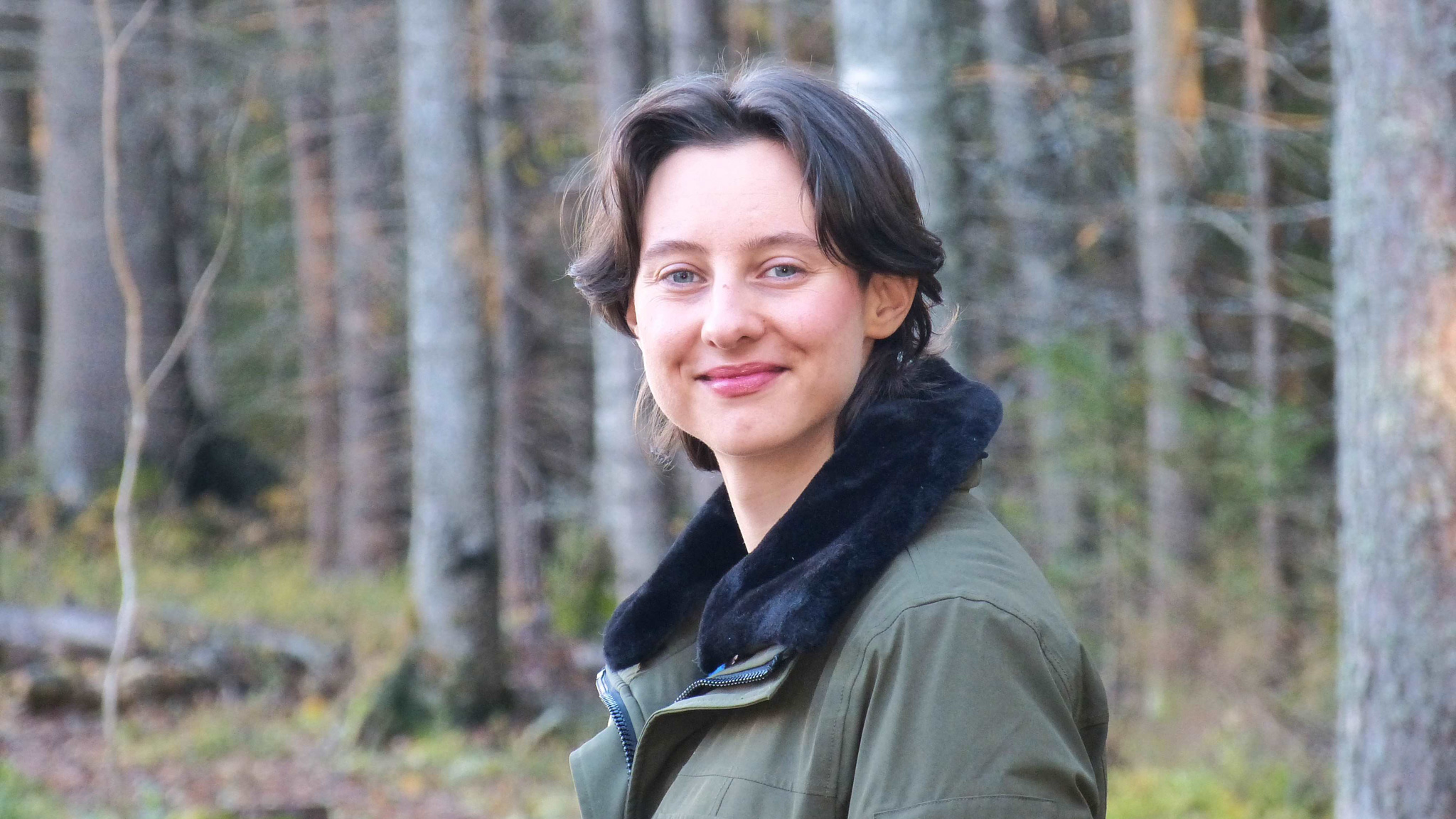 A person with short dark hair and a green jacket stands smiling in a forest with tall trees in the background.