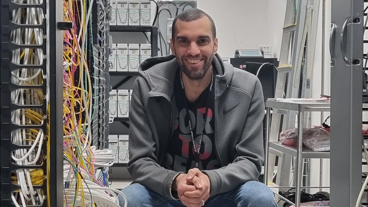 A man sits on a chair in a server hall with a lot of wires and looks happily into the camera