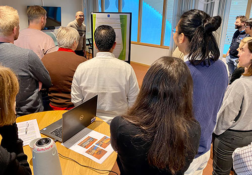 About 20 people stand in a meeting room listening to a man present his scientific poster. Photo.