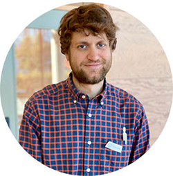 Headshot of a young man with dark brown hair and beard and blue chequered shirt. Photo.