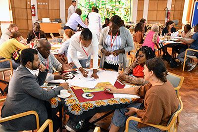 About 20 African women and men, sitting around round tables and discussing. Photo.