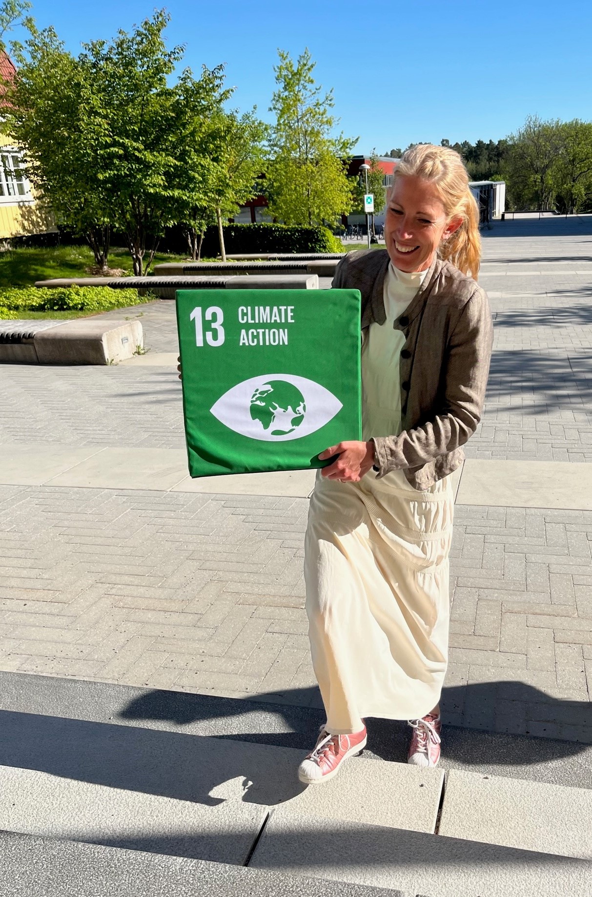 En kvinna i lång ljus kjol och ljusbrun jacka går uppför en trappa hållandes i en grön kub med texten 13 Climate action