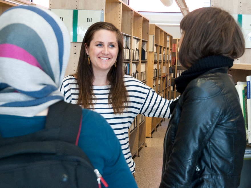 An image of three people standing up, talking. They are in a library, books in the background.