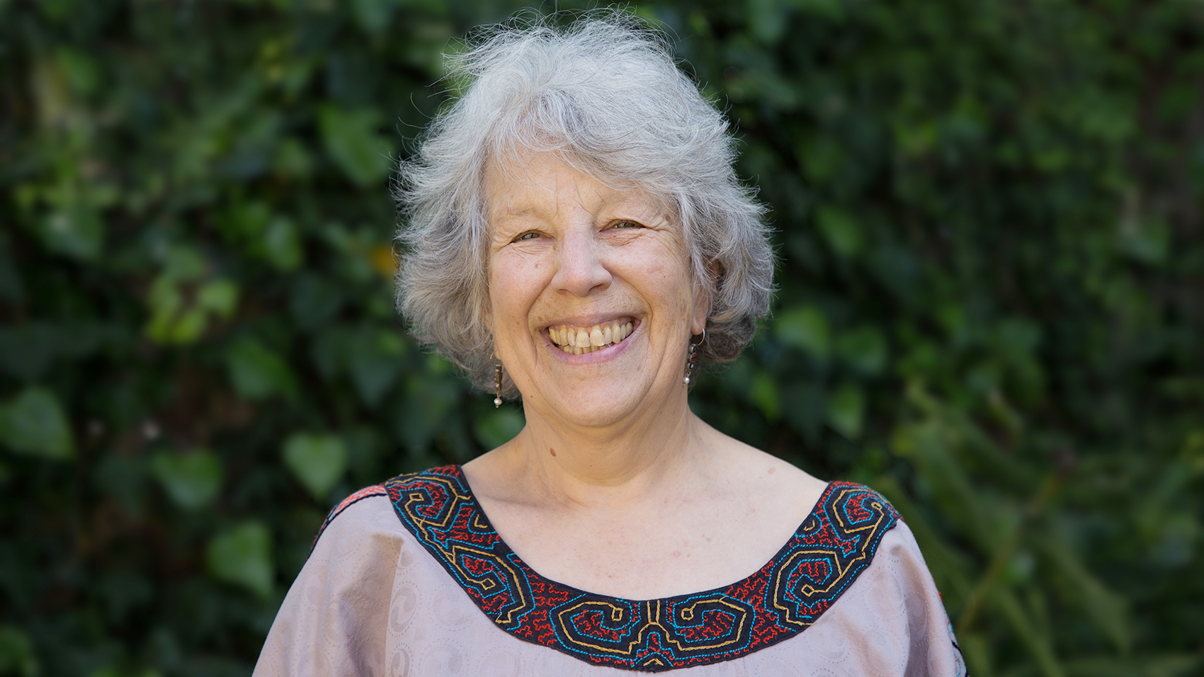 An older woman smiling in front of a bush.