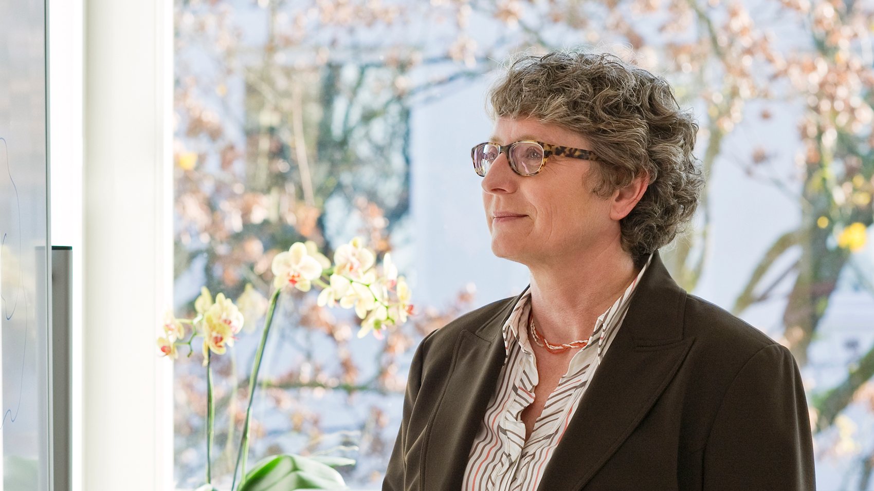 A woman with curly hair wearing glasses standing in front of a window.