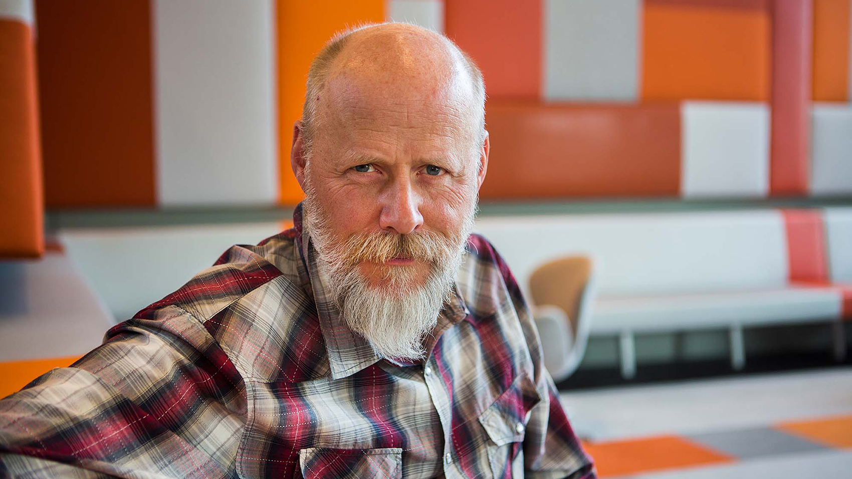 An elderly man with a white beard sitting in a modern indoor environment with orange coloured accents, looking firmly into the camera.
