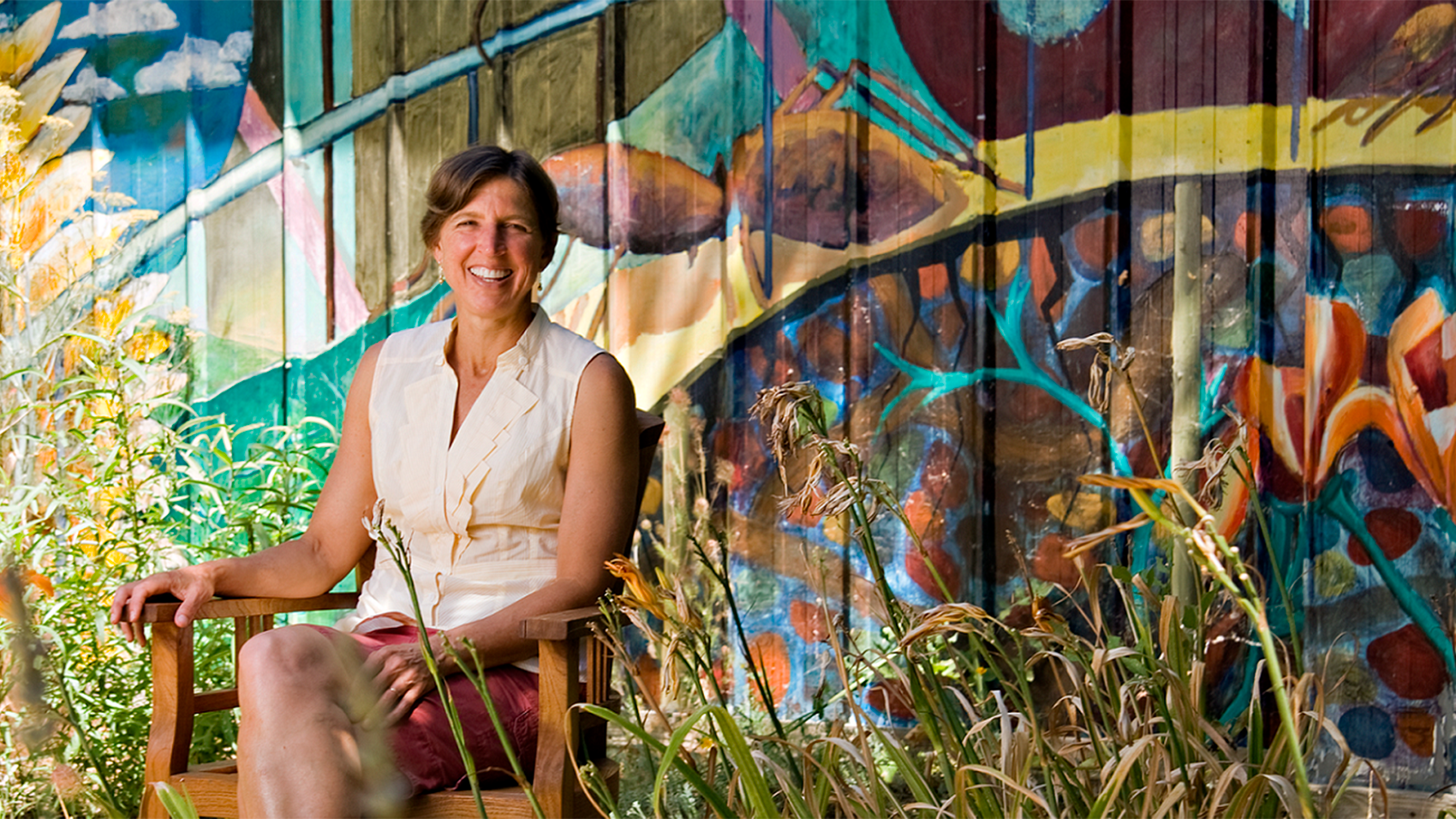 A woman sitting outdoors in a wooden chair in front of a wall with graffiti.