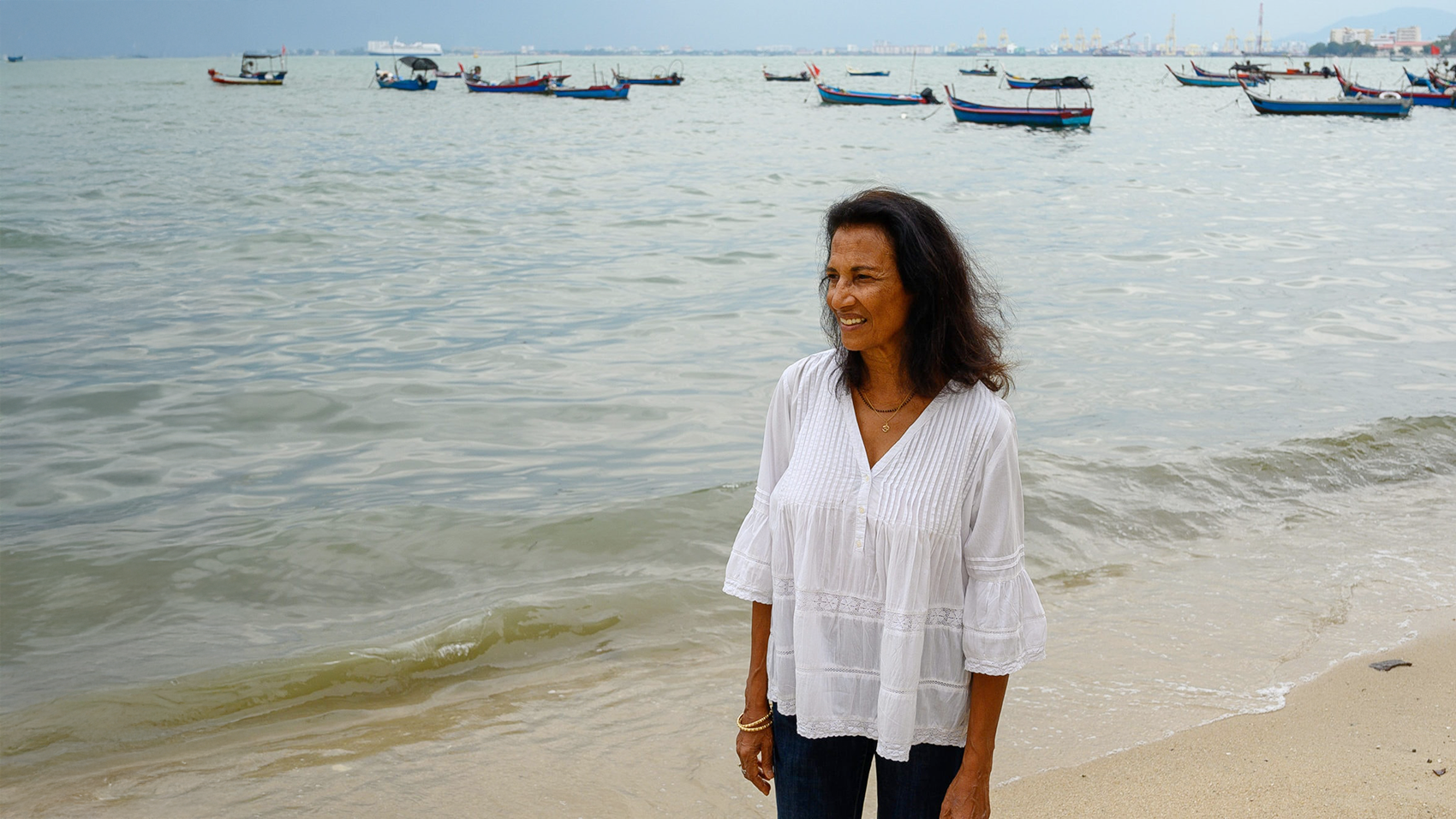 A woman in a white blouse and black hair standing on a beach. Moored small fishing boats are visible behind her.