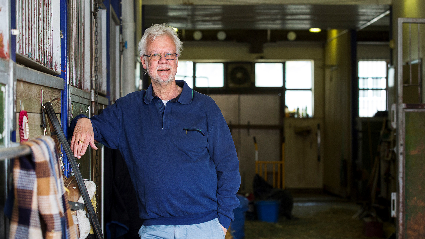 A man in a blue shirt leans himself against the wall in a stable.