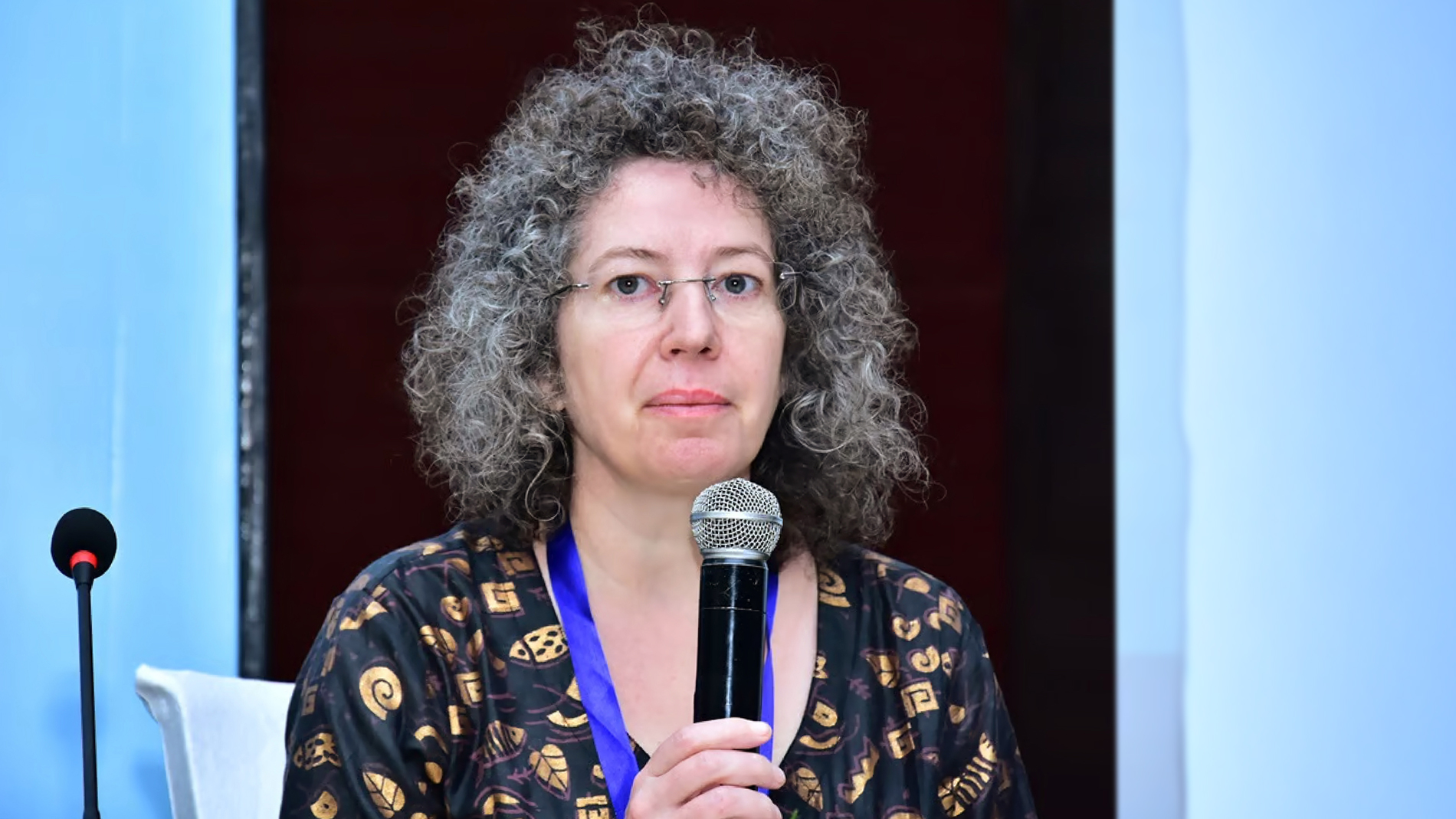 A woman in grey curly hair holds a microphone in front of her.
