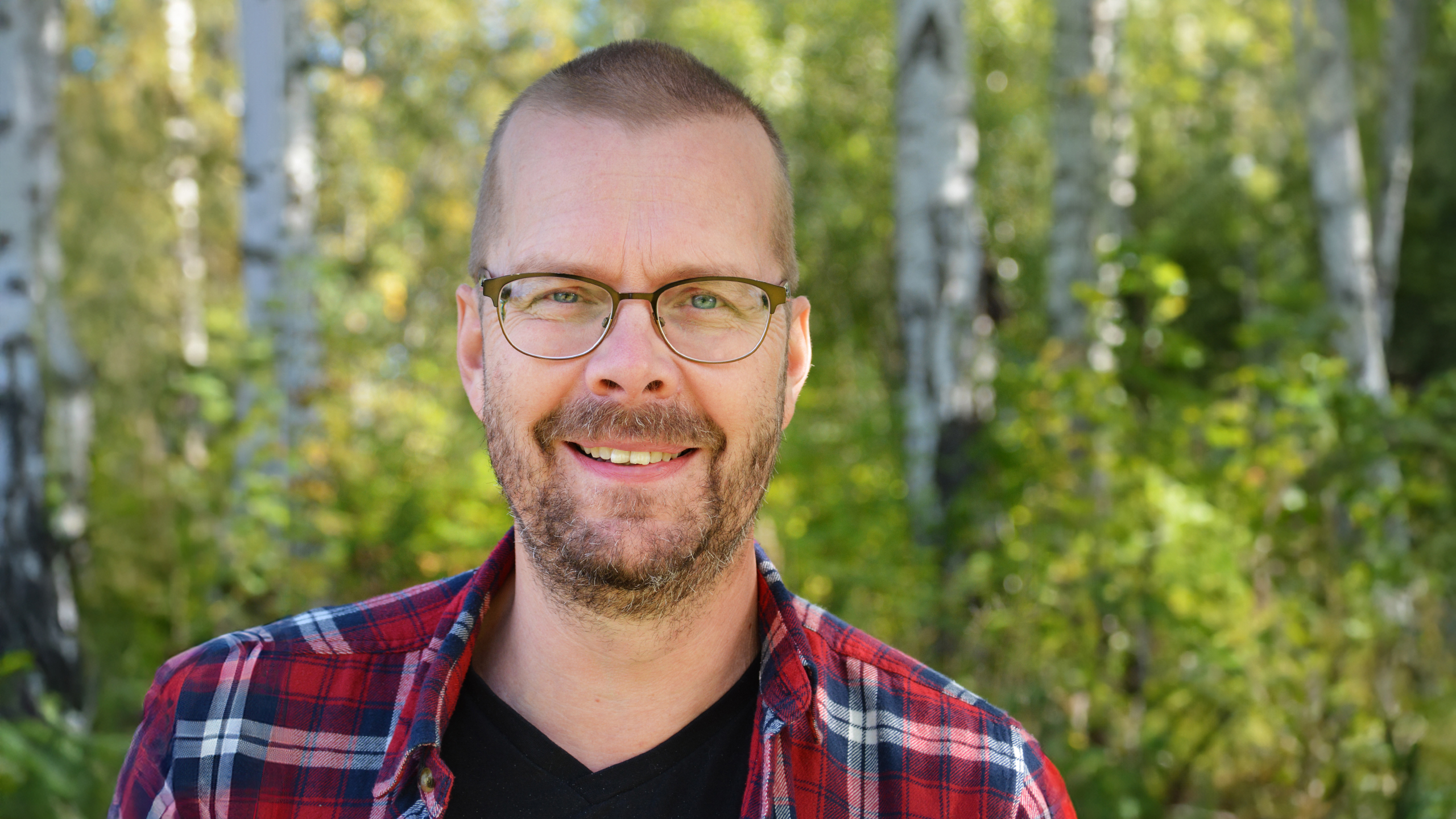 A man with a stubble, wearing glasses and a red checkered shirt, standing in a birch forest.