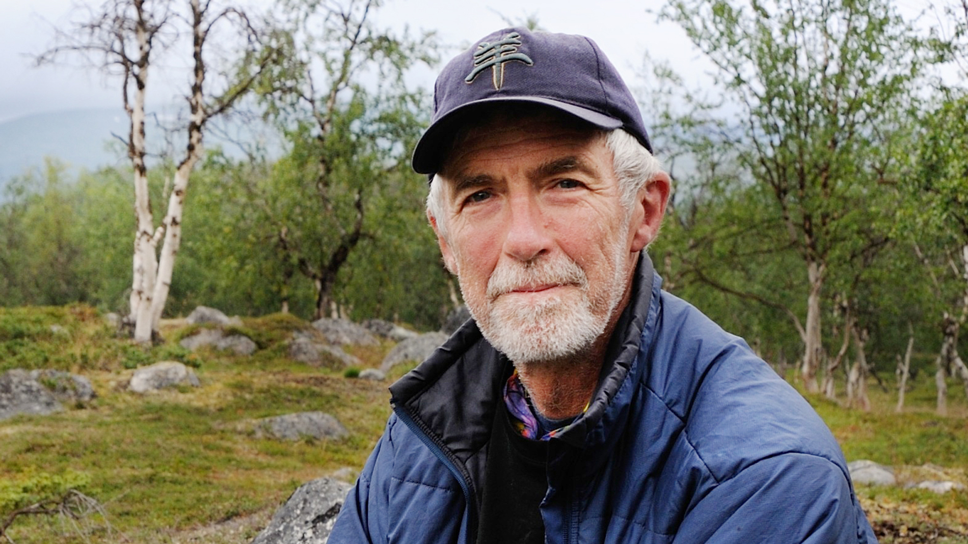 An outdoors dressed man with grey hair and grey beard looks into the camera. In the background a mountain landscape with birch trees with yellow leaves.